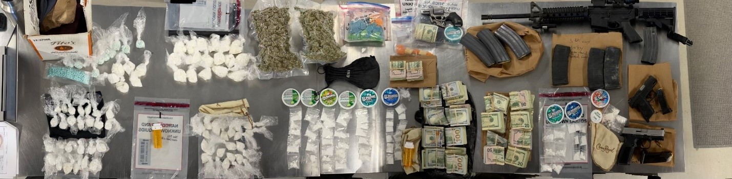 lots of drugs on a table. They are grouped by different size bags. There is also several wads of cash. and an assault rifle and several extra clips. 