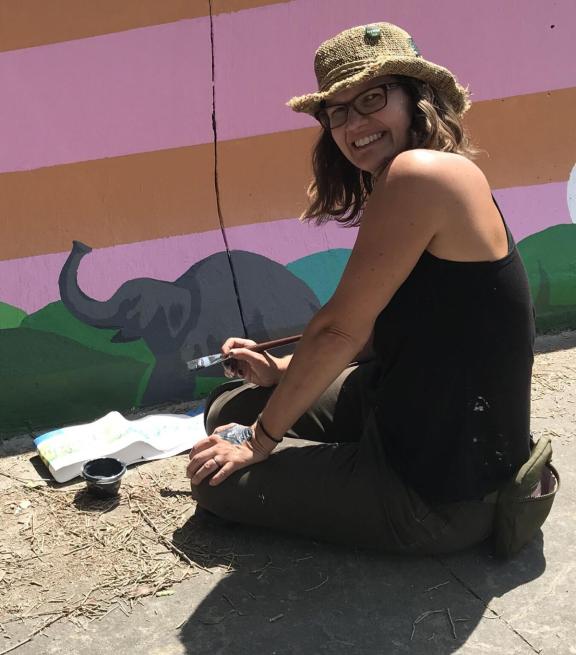kelly kneeling beside her artwork with paintbrush in hand
