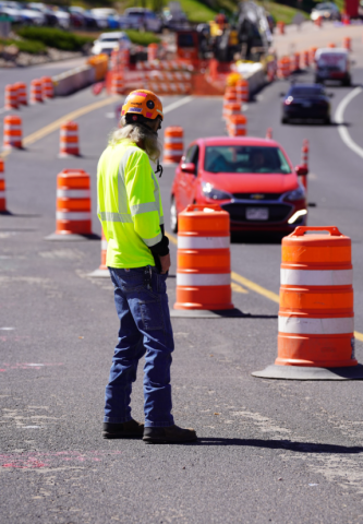 A public works employee in a safety vest stands behind orange cones while cars pass on the road nearby.