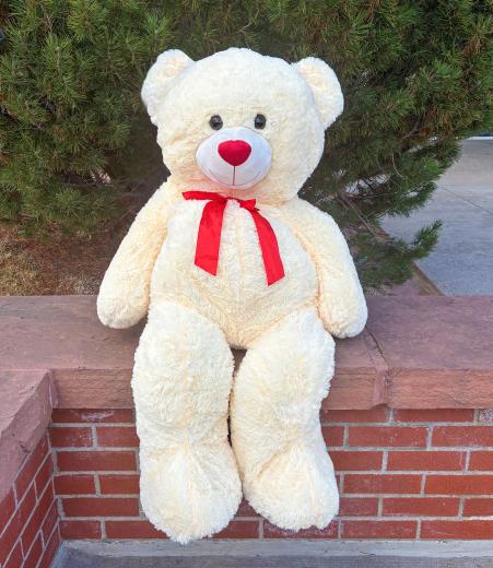 A large cream colored teddy bear with a red bow sitting on a brick wall.