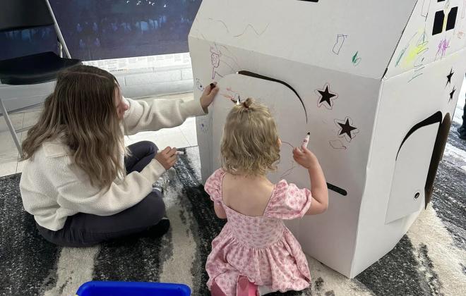 a little girl colors a large cardboard house with the help of a teacher
