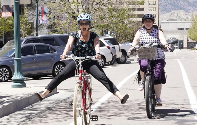 two women riding bikes downtown. One has her feet off the pedals and extended to the side in a playful manner. The other is following behind smiling. 