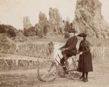 woman stands next to a man on a bike. Large rocks in the background.