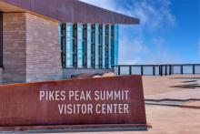 Pike Peak Summit Visitor Center sign. Outdoor viewing platform and wall of windows on the visitor center in the background.